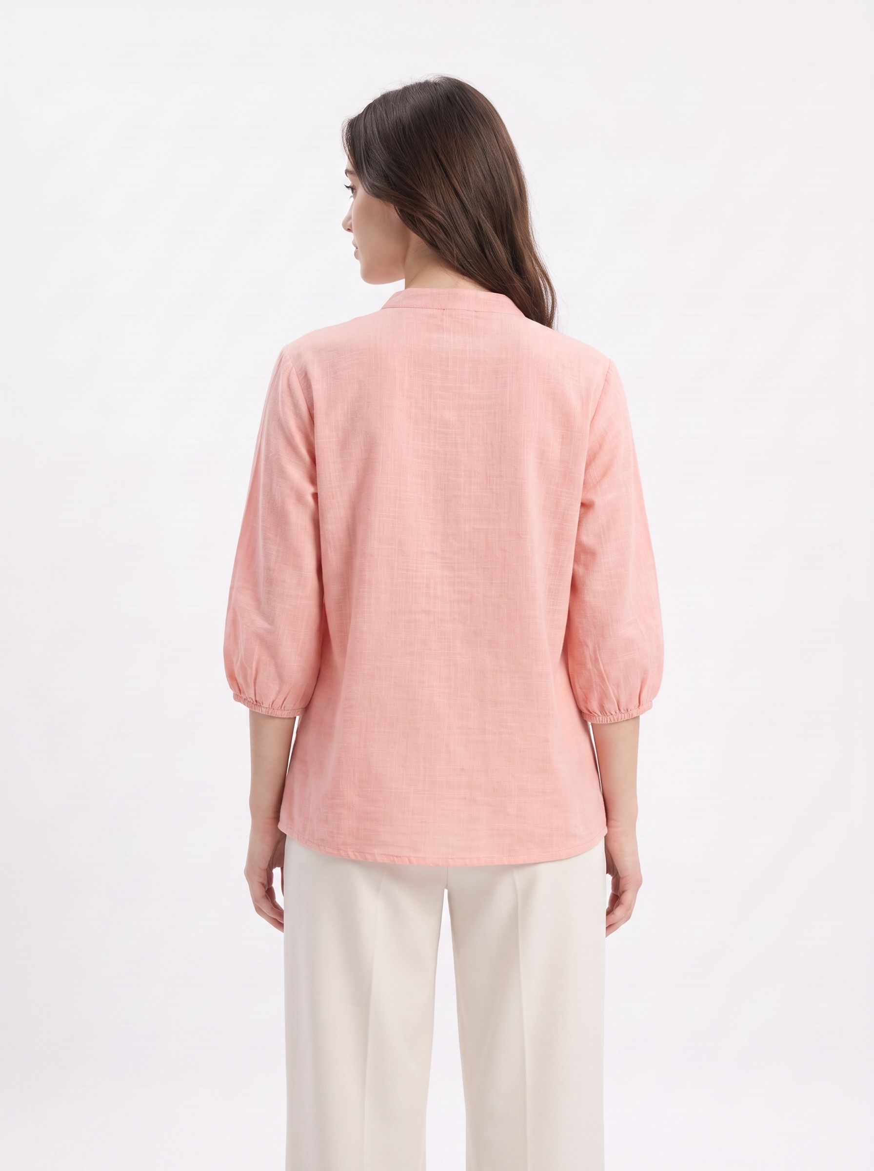 A woman viewed from the back wearing a light pink linen blouse with three-quarter puff sleeves and cream-colored trousers, set against a plain white studio background.