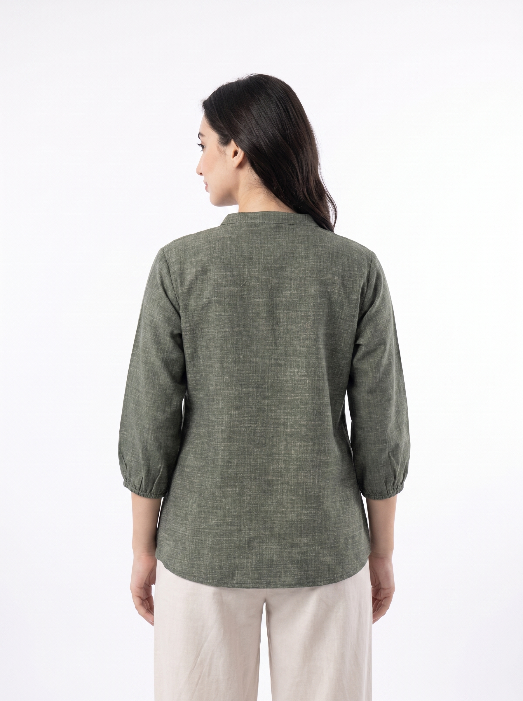 A woman viewed from the back wearing a textured olive green three-quarter sleeve top and light beige trousers, set against a plain white studio background.