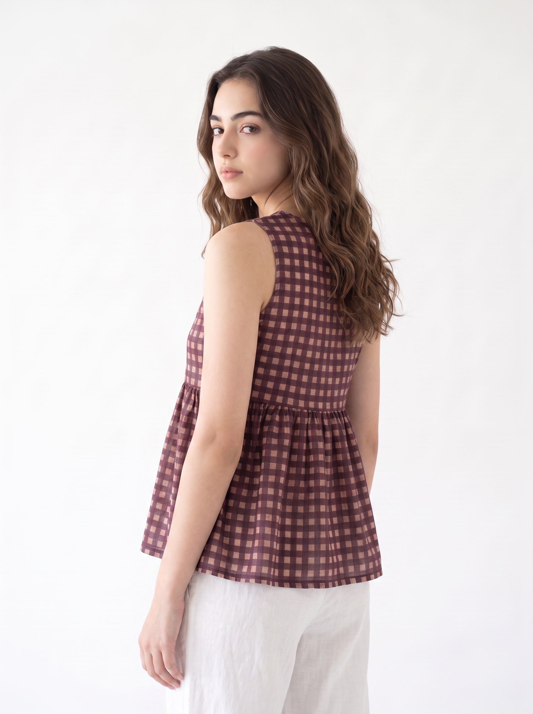 A woman wearing a sleeveless peplum top with a brown and pink gingham pattern, paired with white pants, photographed against a plain white studio background.