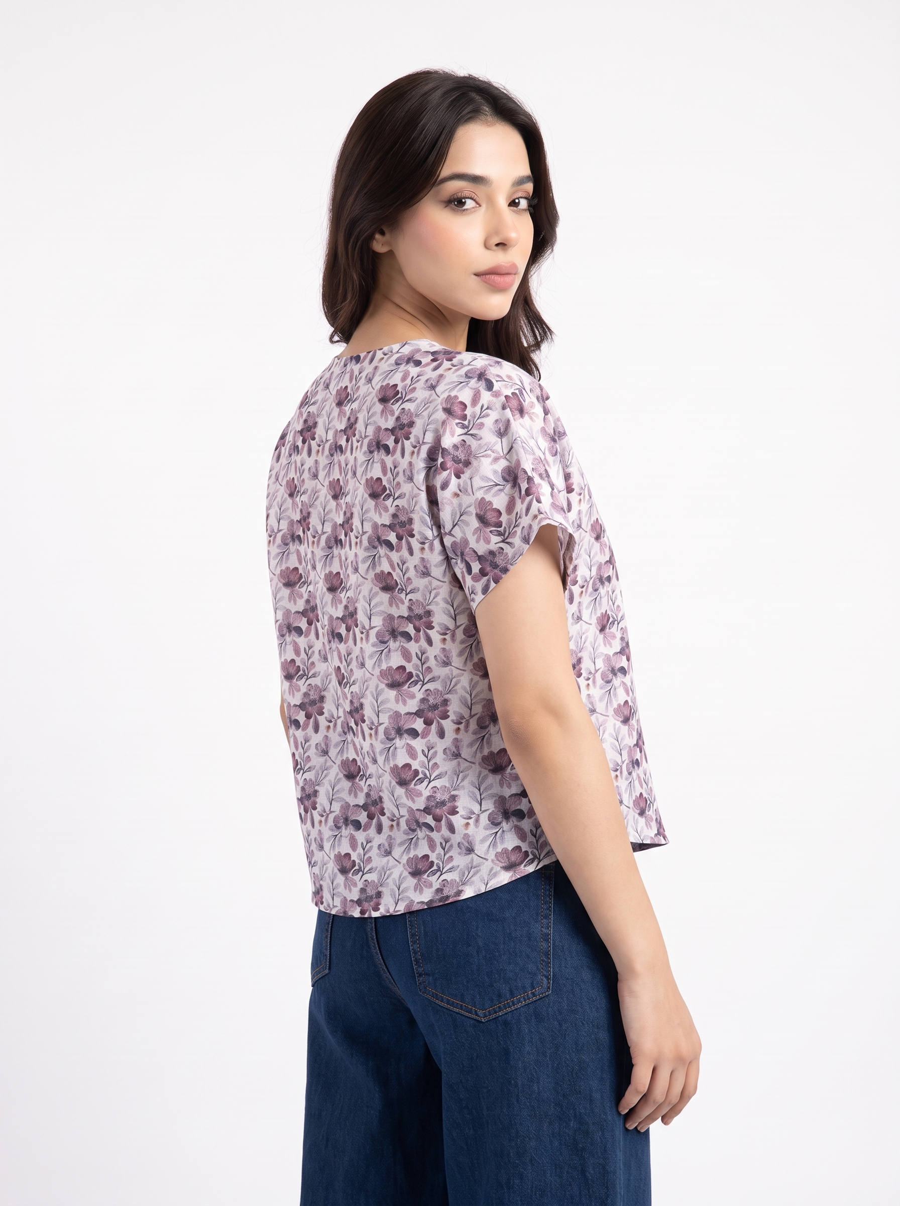 A woman shown from the back wearing a short-sleeve cropped top with a muted purple and gray floral pattern on a light background, paired with dark blue jeans, against a plain white studio background.