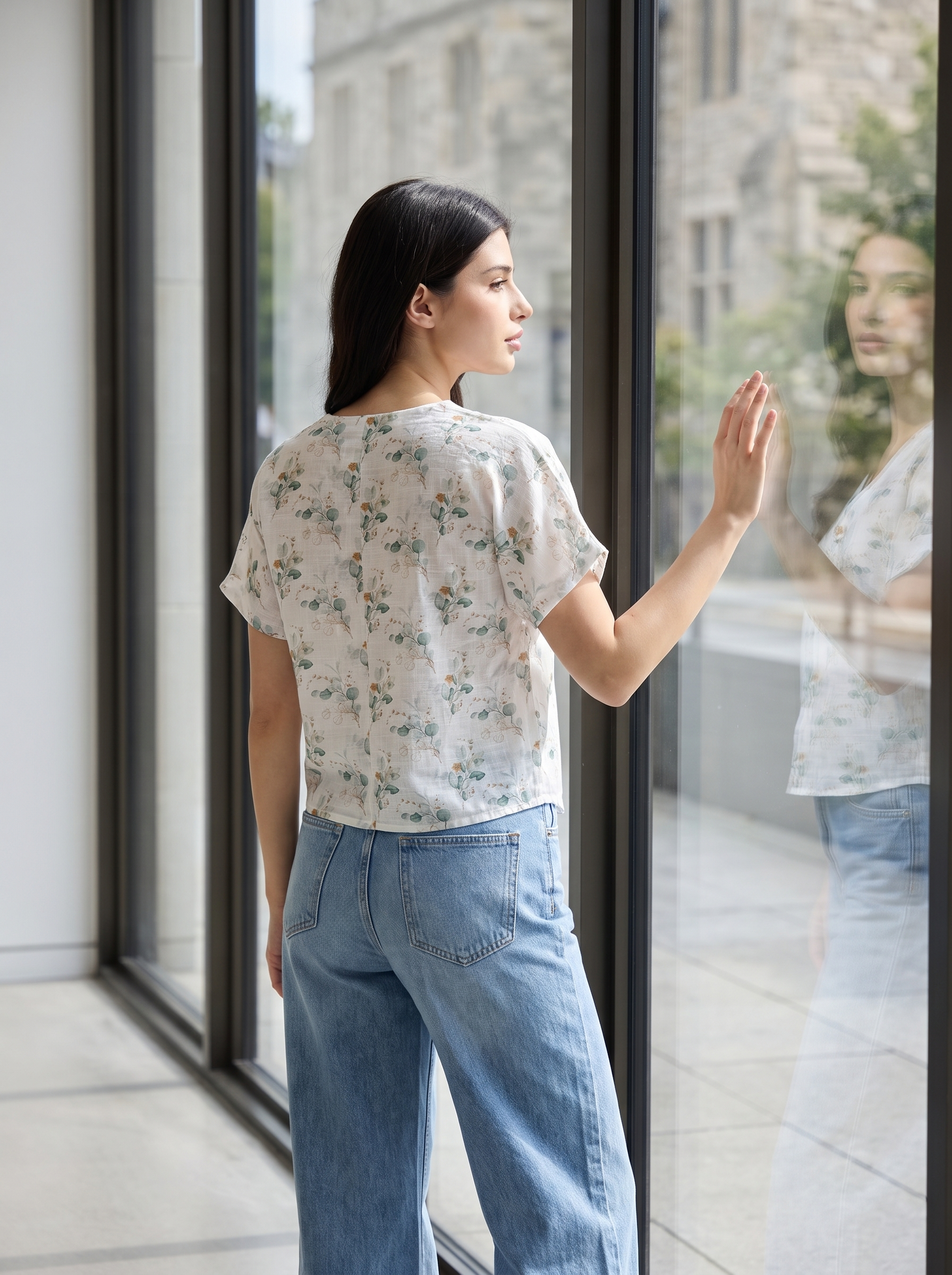 A woman stands by a large glass window, viewed from behind and side, wearing a short-sleeve cream-colored top with a delicate green and blue floral pattern and light blue wide-leg jeans.