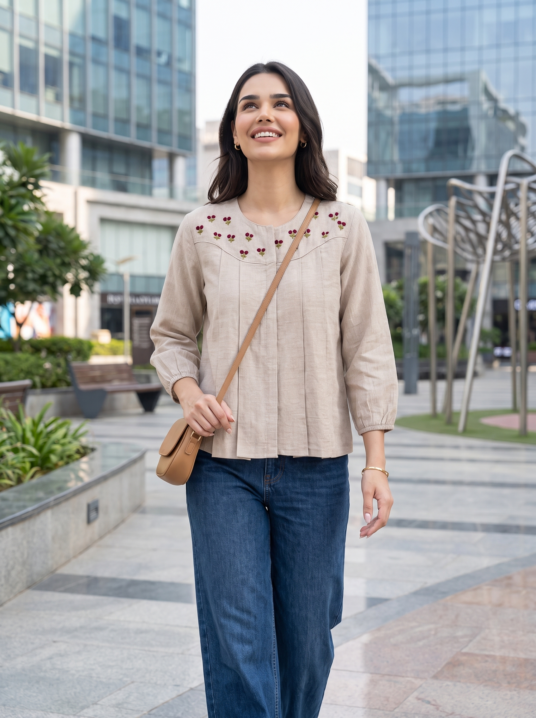 A woman wearing a beige long-sleeve blouse with embroidered details on the chest and blue jeans, carrying a tan crossbody bag, standing in an outdoor urban plaza with modern glass buildings in the background.