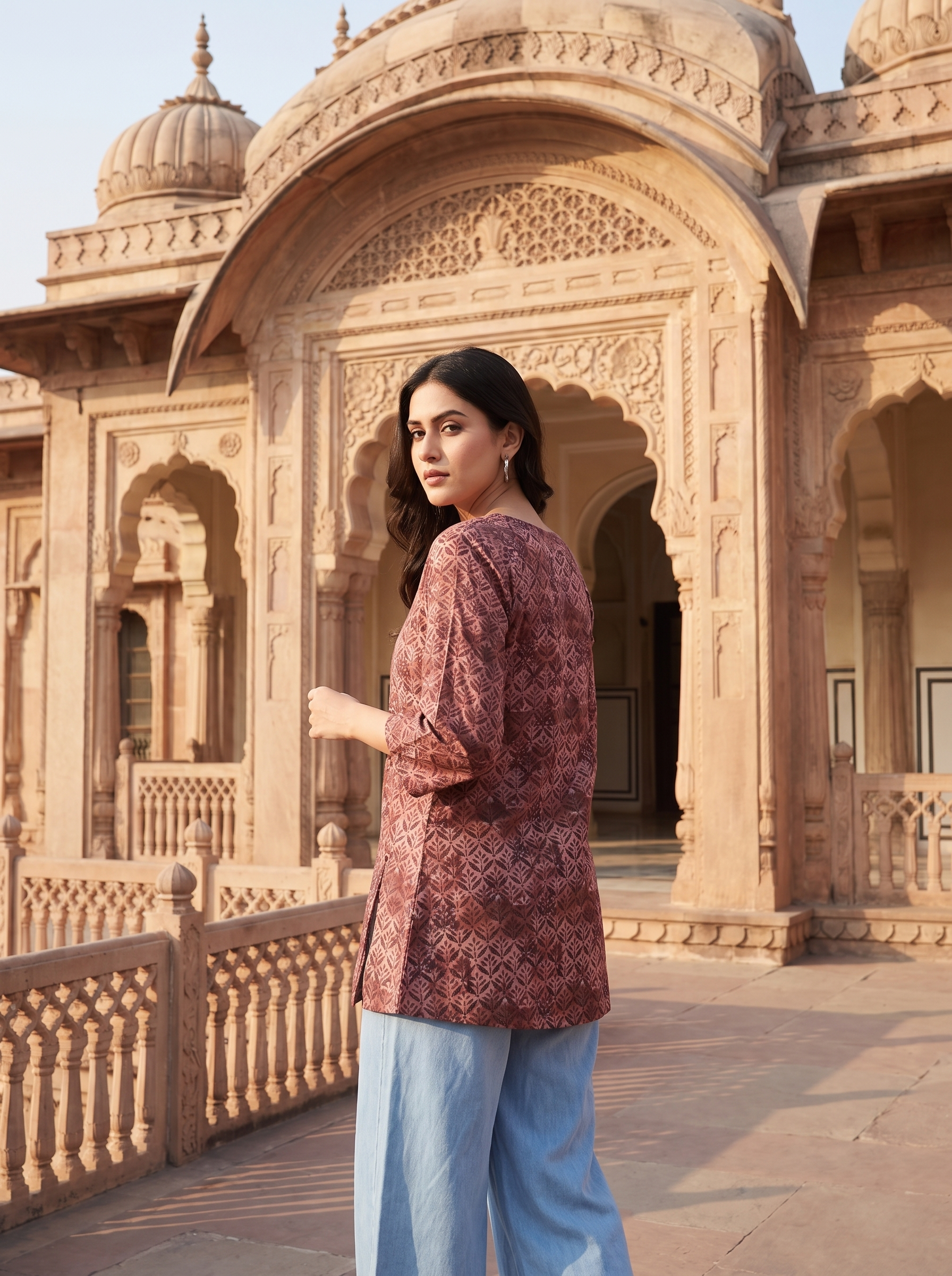 A woman wearing a dusty rose printed kurta and light blue wide-leg trousers stands in front of a historic sandstone palace with intricate arches and domes.