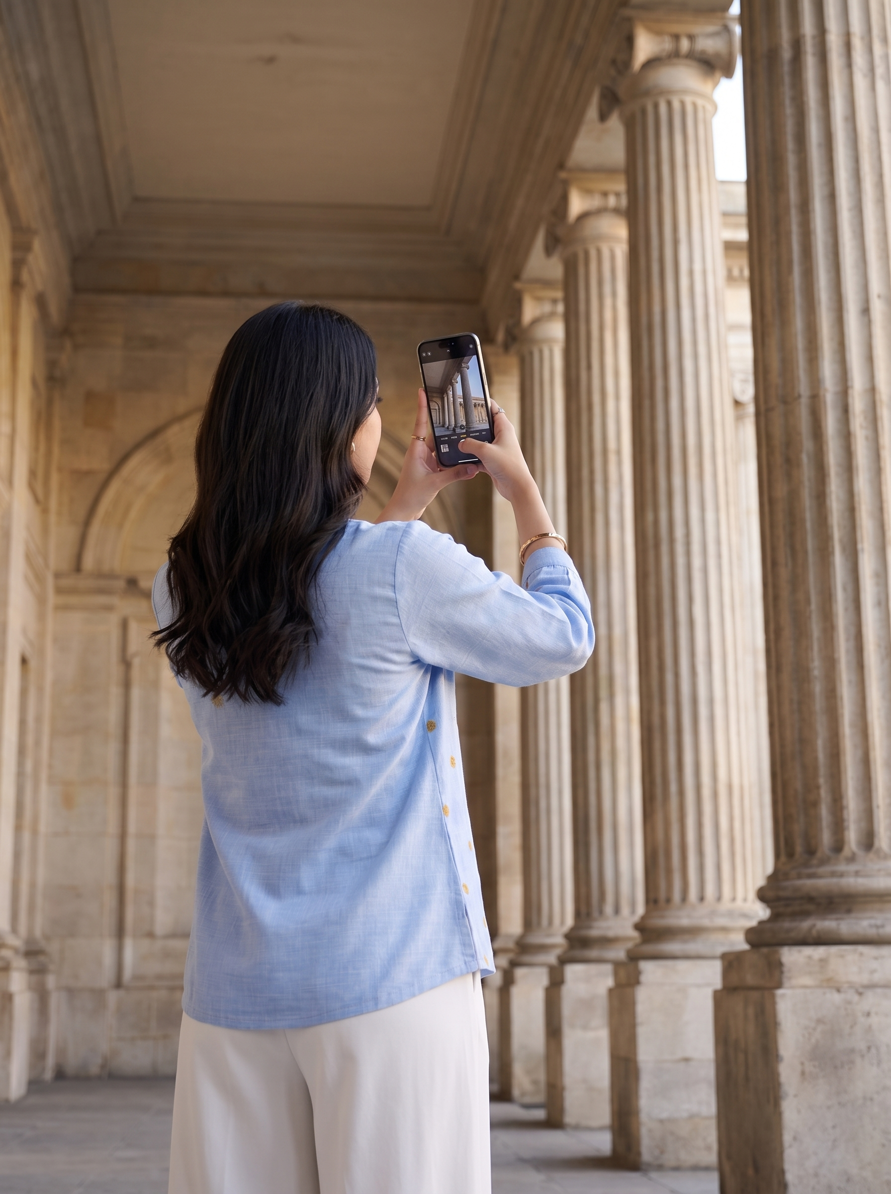 A woman with long dark hair stands in a stone colonnade, photographed from behind as she holds up a smartphone to take a photo. She is wearing a light blue button-front top and cream-colored pants.