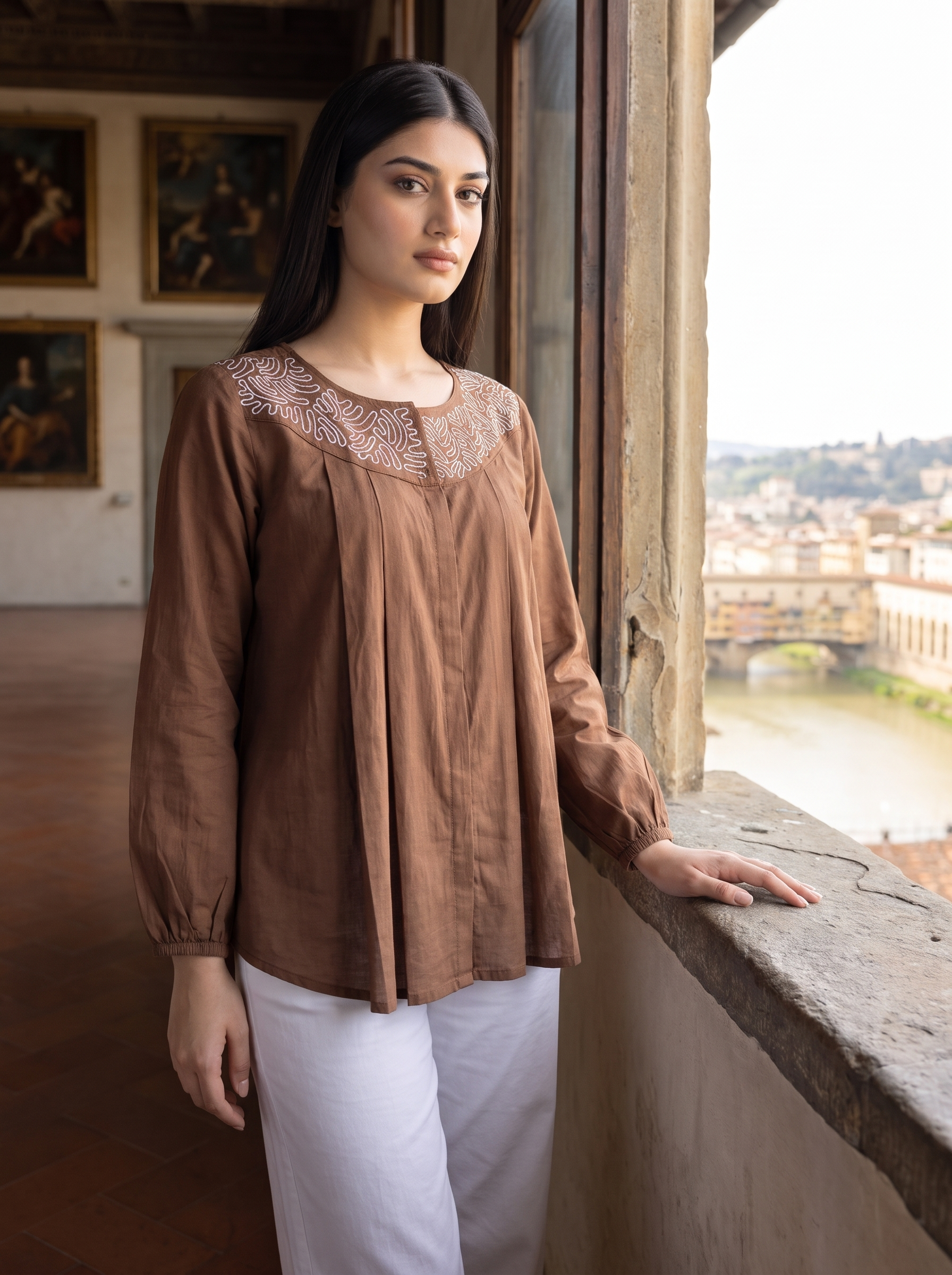 A woman wearing a brown long-sleeve linen blouse with a detailed embroidered neckline and white pants, standing by a window overlooking a scenic city view.