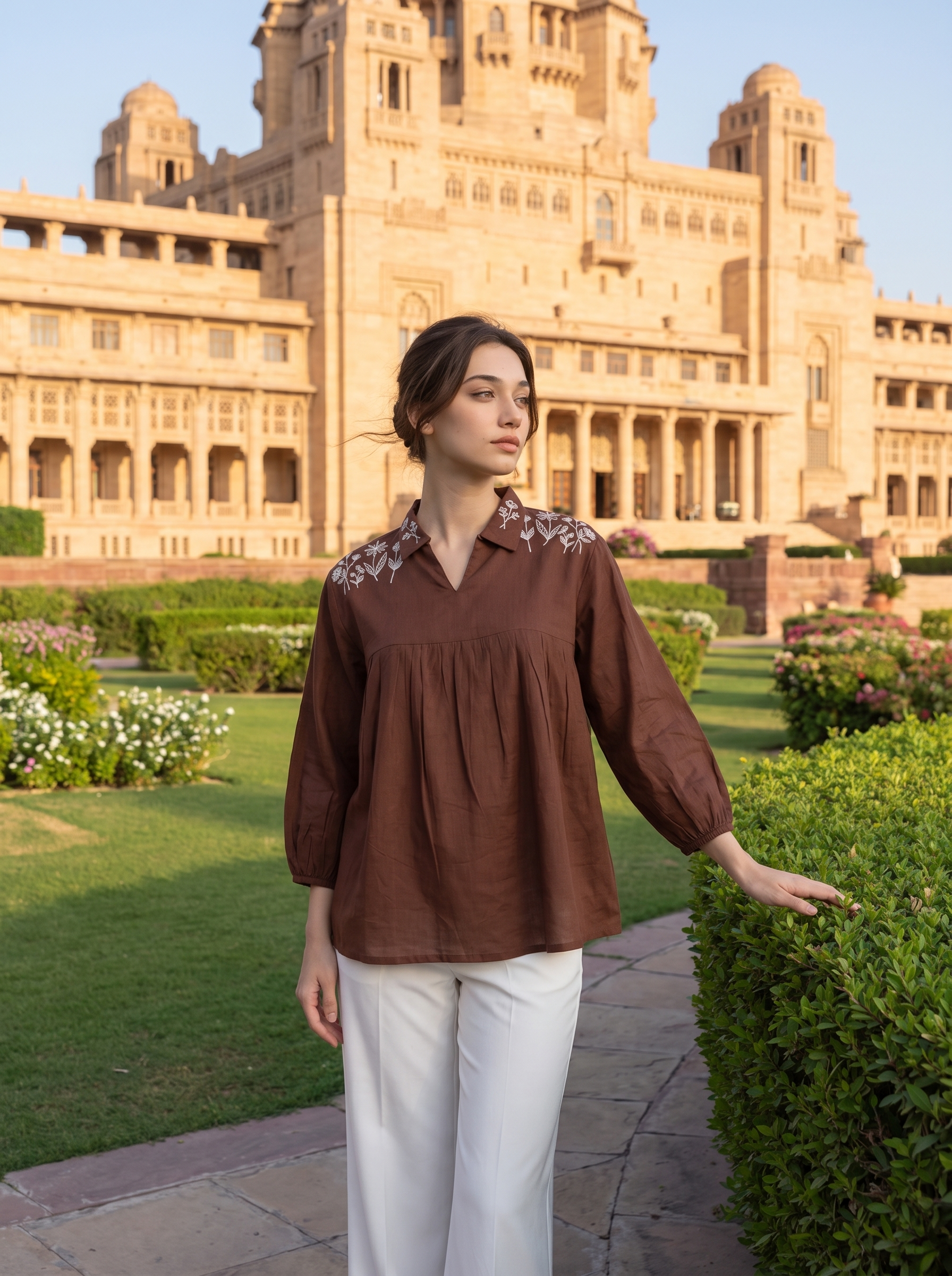 A woman stands outdoors in front of a large sandstone palace and manicured gardens, wearing a brown long-sleeve blouse with embroidered detailing on the shoulders and white pants.