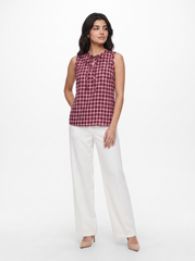 A woman modeling a sleeveless red and black checkered blouse with a tie-neck detail, paired with white wide-leg trousers and beige heels, against a plain white studio background.