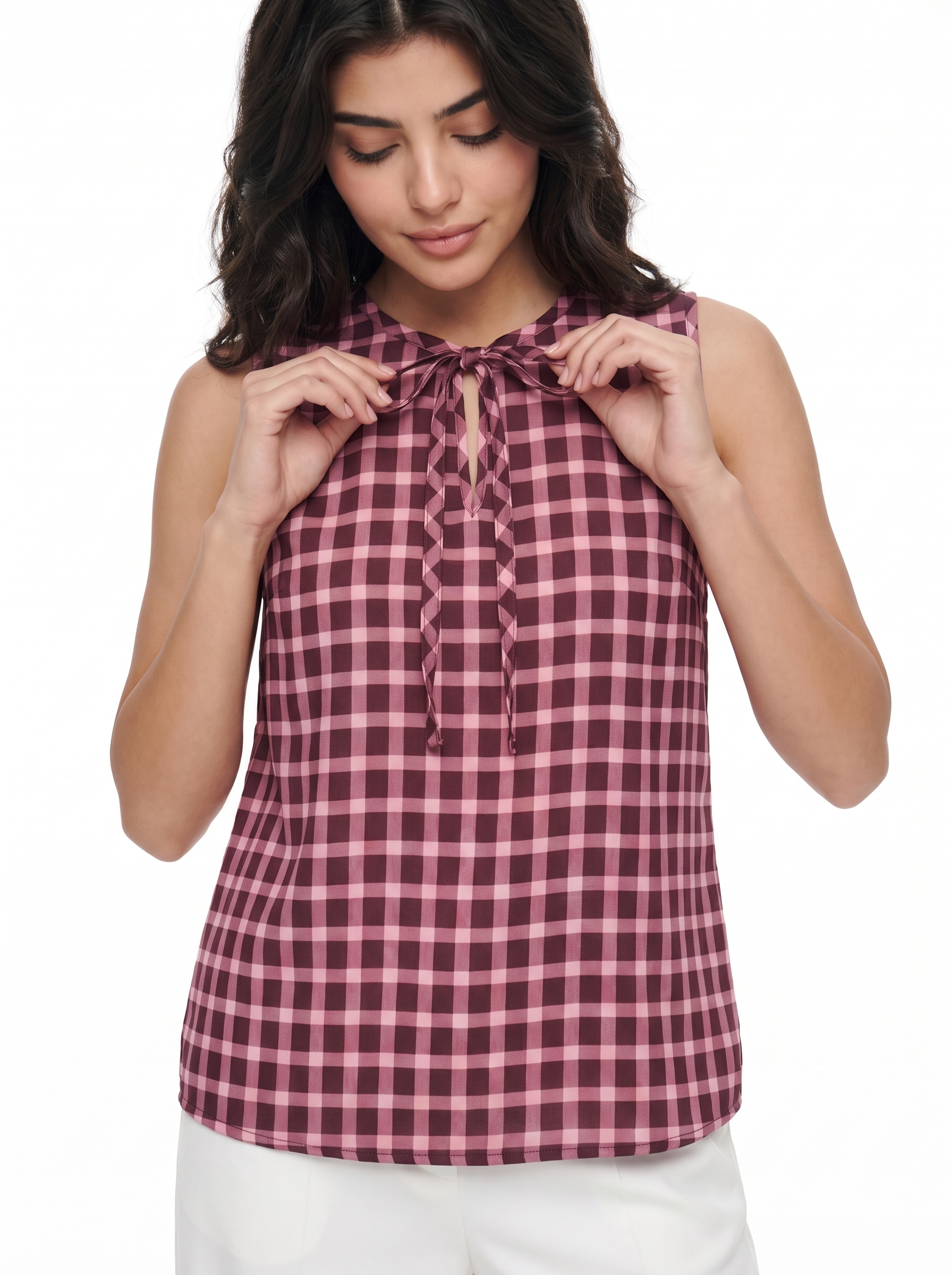 A woman wearing a sleeveless gingham plaid top in dusty pink and burgundy with a tie-neck detail, paired with white pants, against a solid white background.