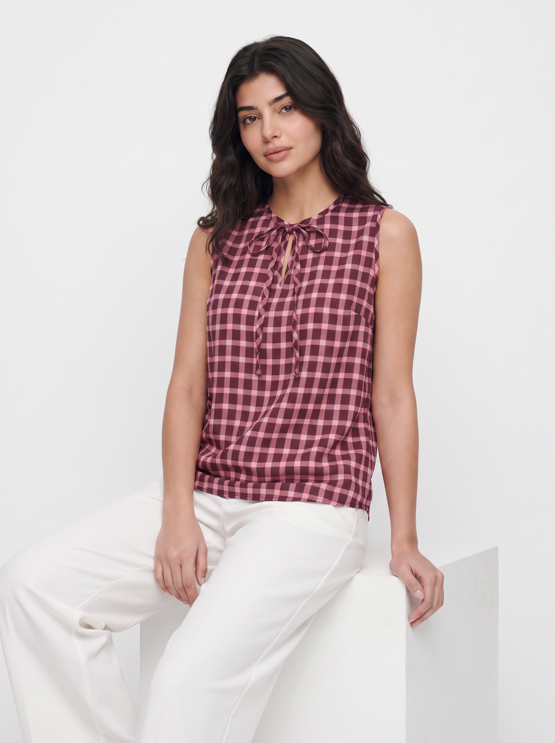 A woman wearing a sleeveless red and black gingham tie-neck blouse with white pants, sitting on a white block against a white studio background.