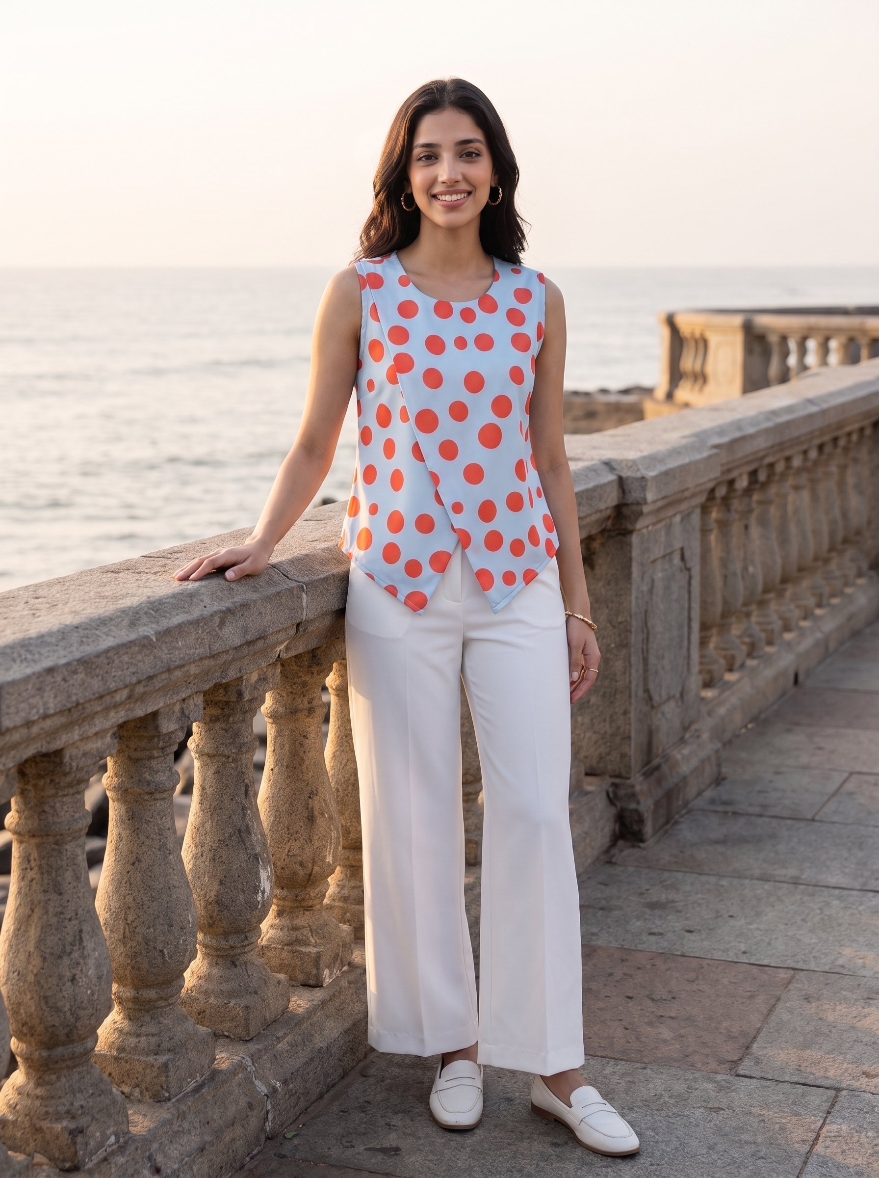 A woman stands on a stone walkway by the sea, wearing a sleeveless wrap-style top with orange polka dots on a light blue background and white wide-leg pants.