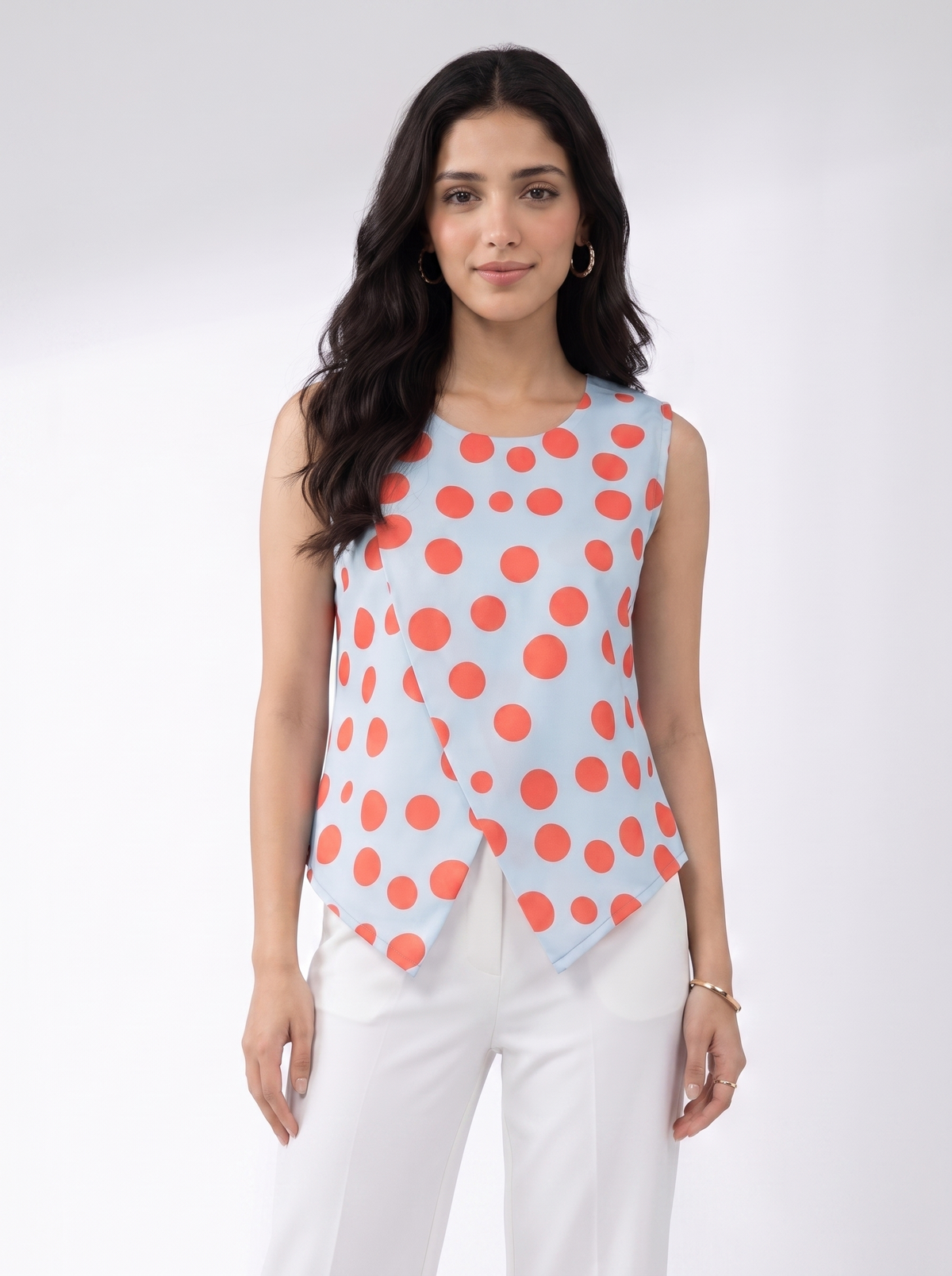 A woman modeling a sleeveless wrap-style top with a light blue base and large coral-red polka dots, paired with white pants, against a plain light gray studio background.