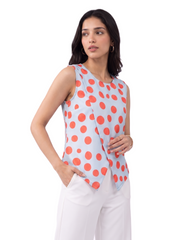 A woman modeling a sleeveless wrap-style top with a light blue base and large coral-orange polka dots, paired with white pants, against a plain white studio background.