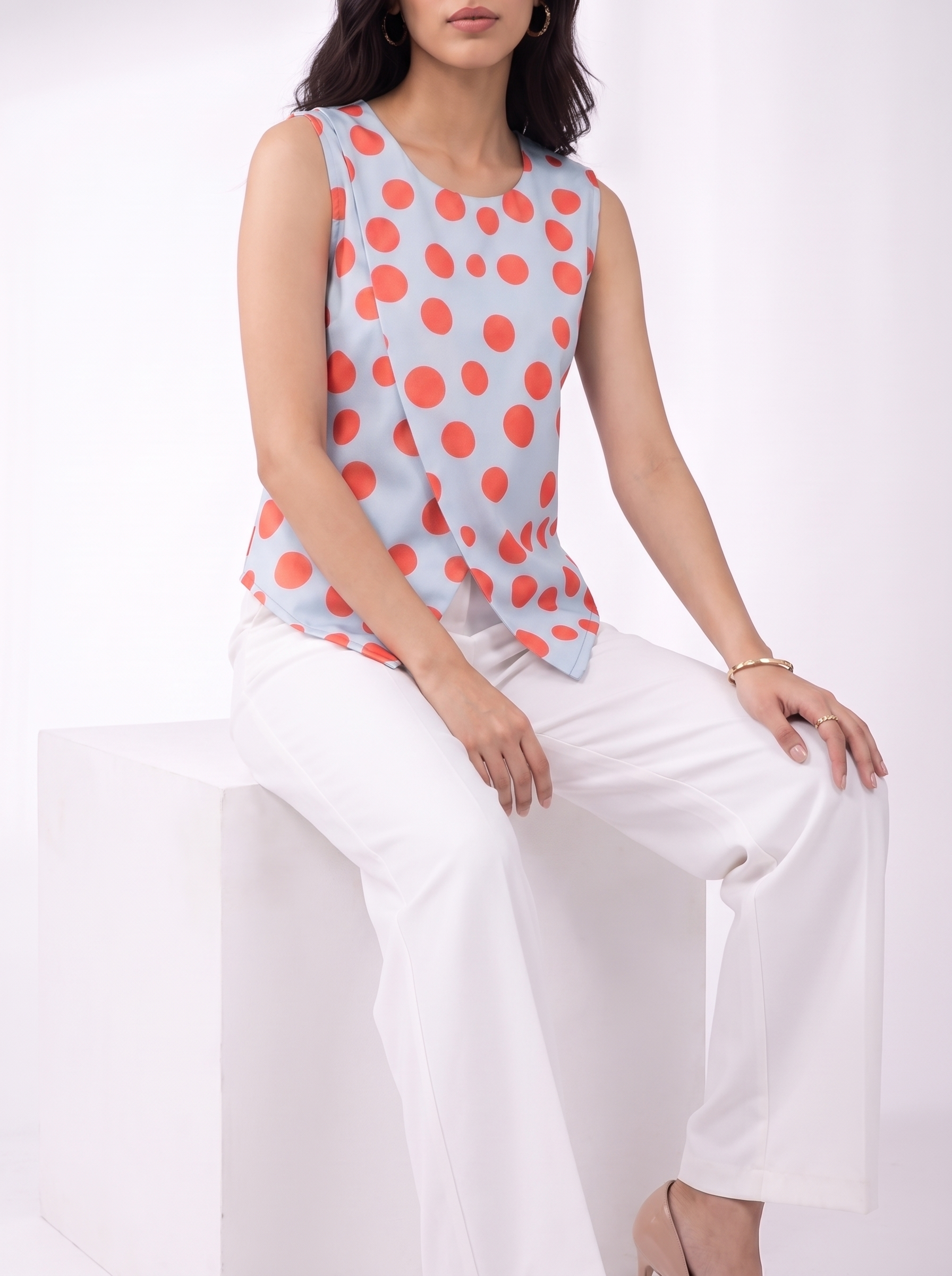 A woman seated on a white block wearing a sleeveless light blue top with large coral-orange polka dots and white wide-leg pants against a clean white studio background.