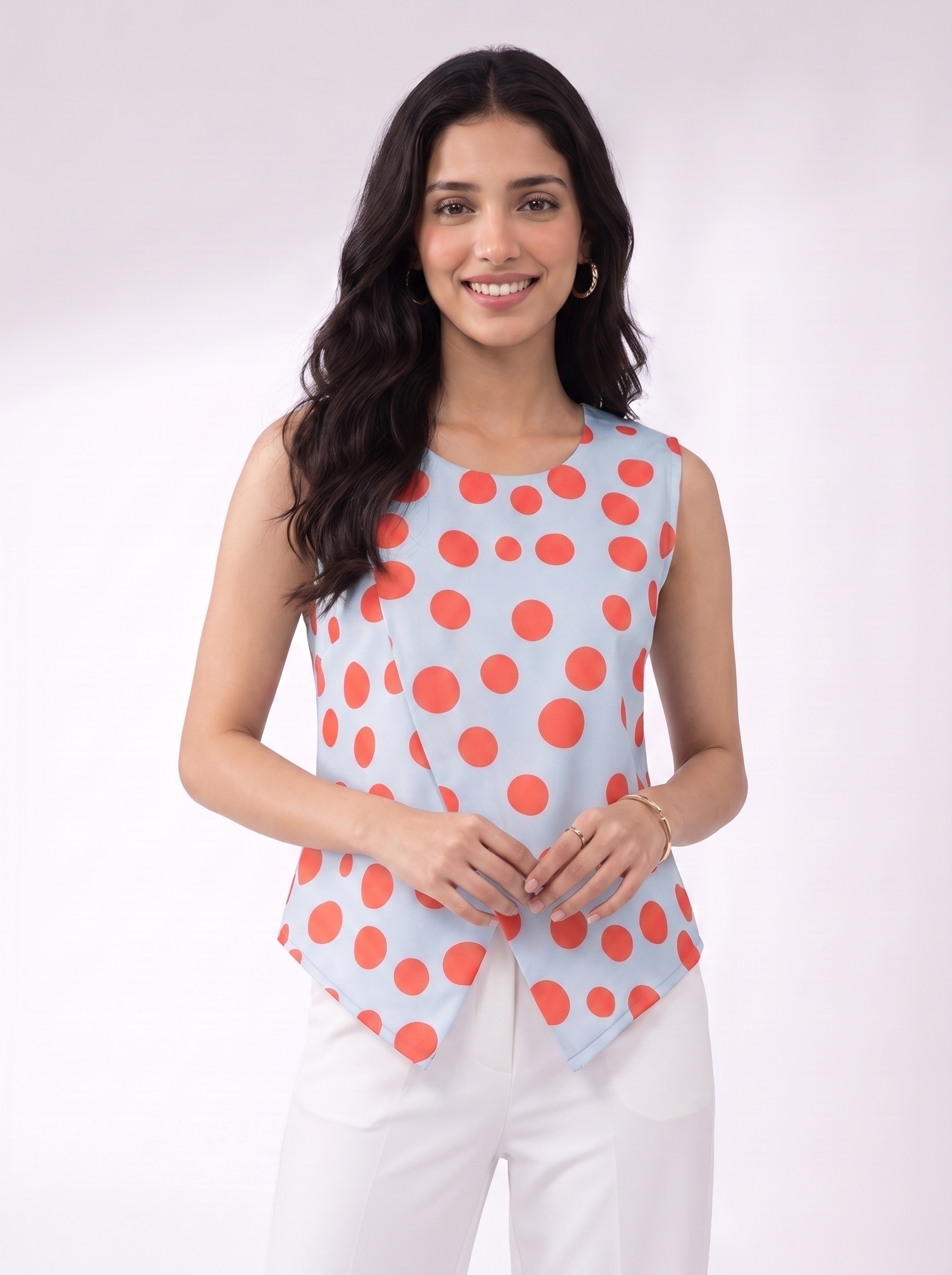 A woman modeling a sleeveless light blue top with a bold coral-red polka dot pattern, paired with white pants, against a clean white studio background.