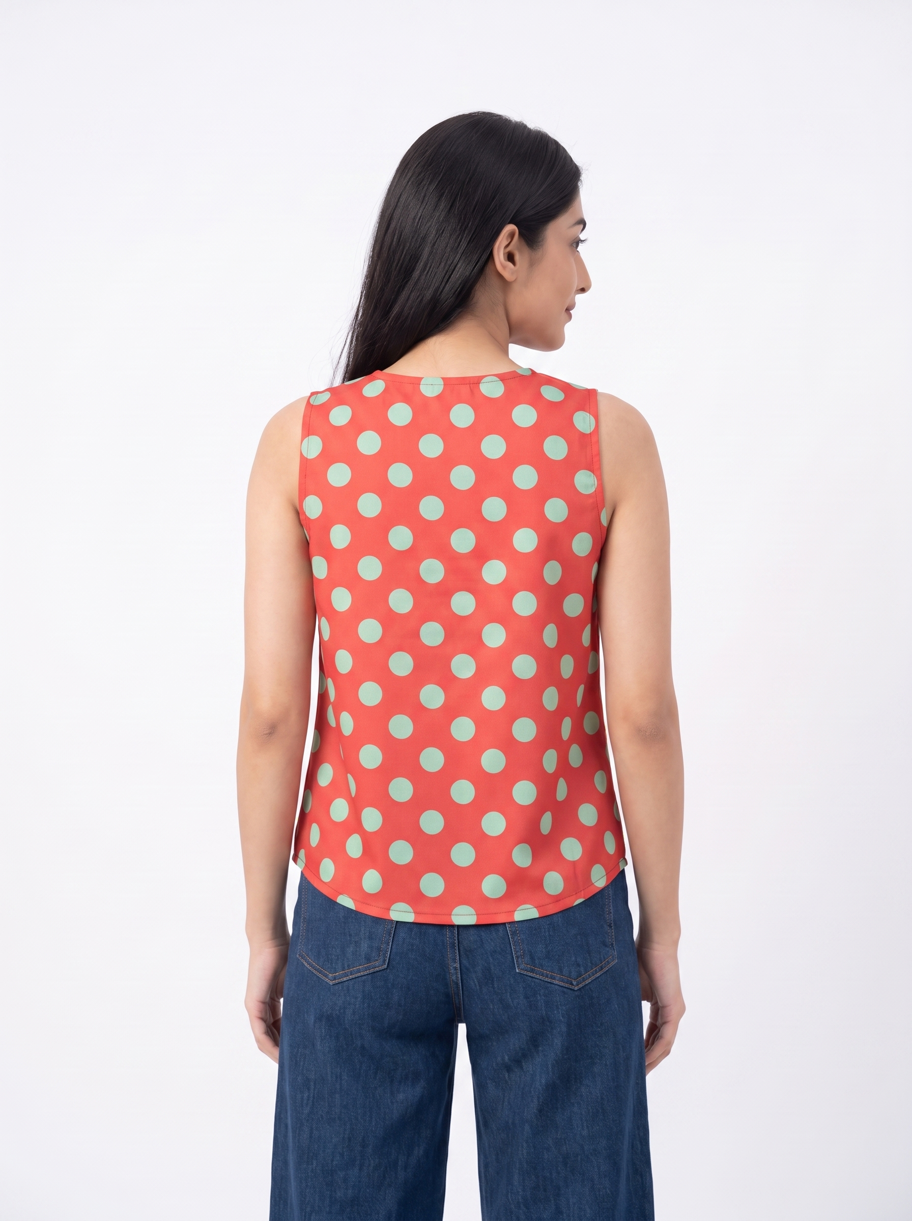 A woman shown from the back wearing a sleeveless coral-red top with a mint green polka dot pattern, paired with dark blue jeans, against a plain white studio background.