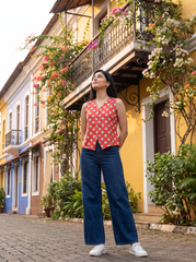 A woman stands on a cobblestone street in front of colorful colonial-style buildings, wearing a sleeveless coral-red top with white polka dots and dark blue wide-leg jeans.