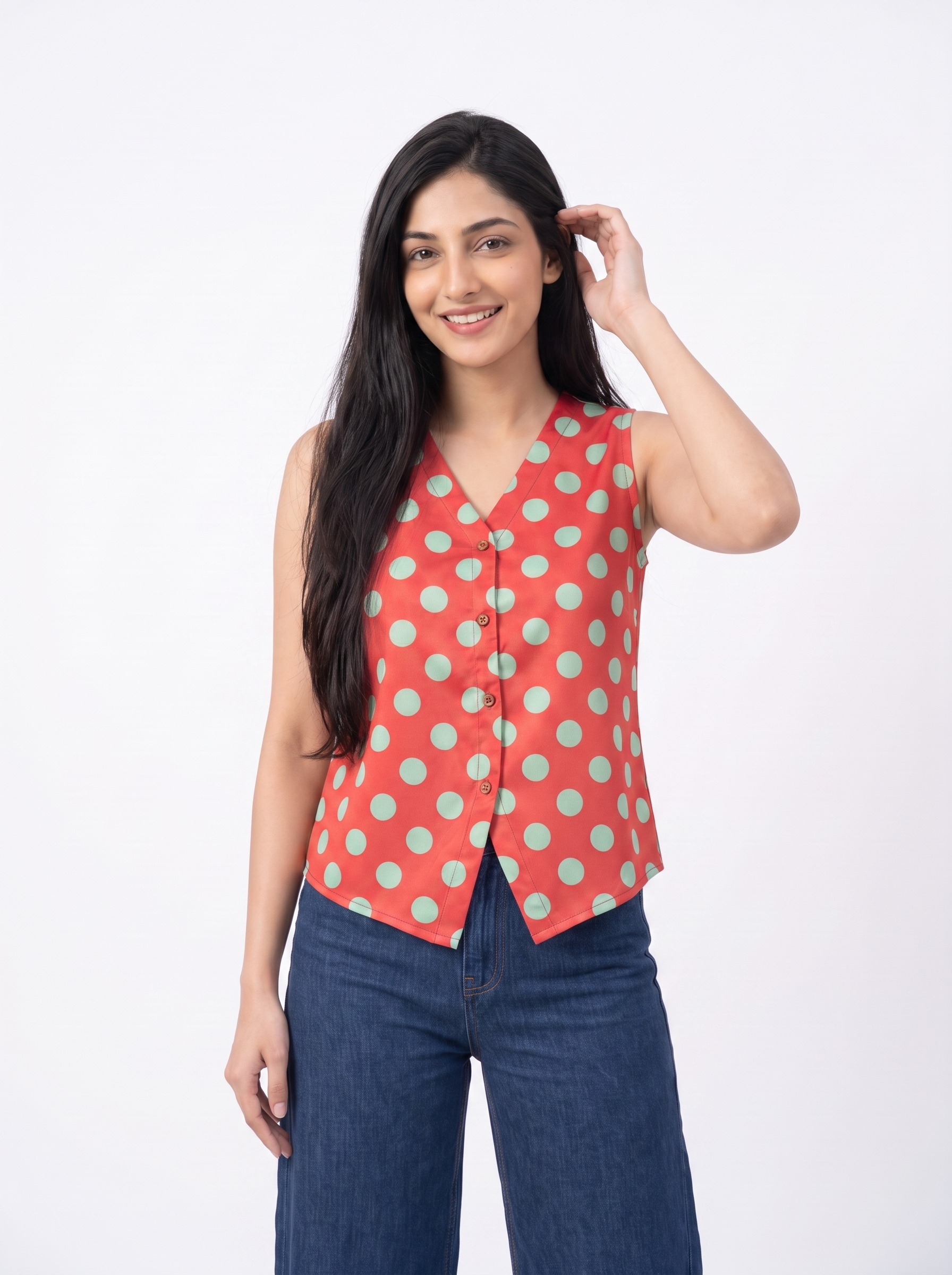 A woman modeling a sleeveless red vest with a mint green polka dot pattern, paired with blue jeans, against a plain white studio background.