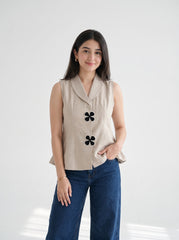 A woman modeling a sleeveless beige linen vest with black four-petal embellishments, paired with dark blue jeans, against a bright white studio background.