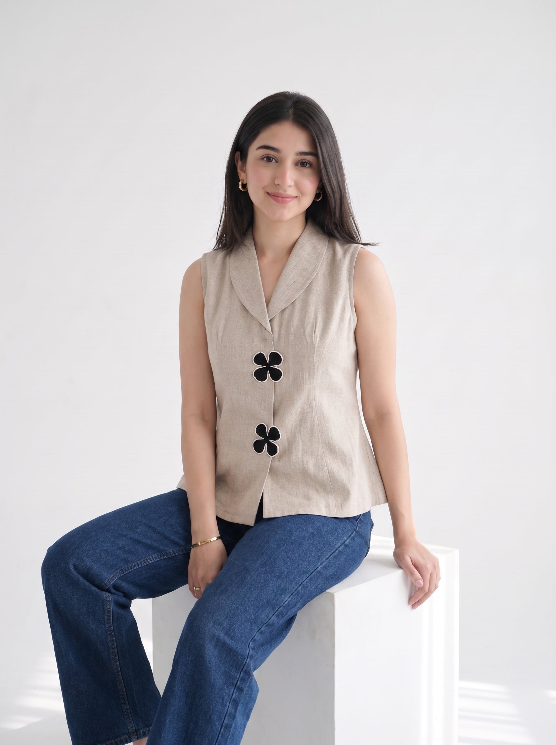 A woman sits on a white block pedestal wearing a sleeveless beige linen vest with black flower-shaped buttons and blue jeans, set against a clean white studio background.