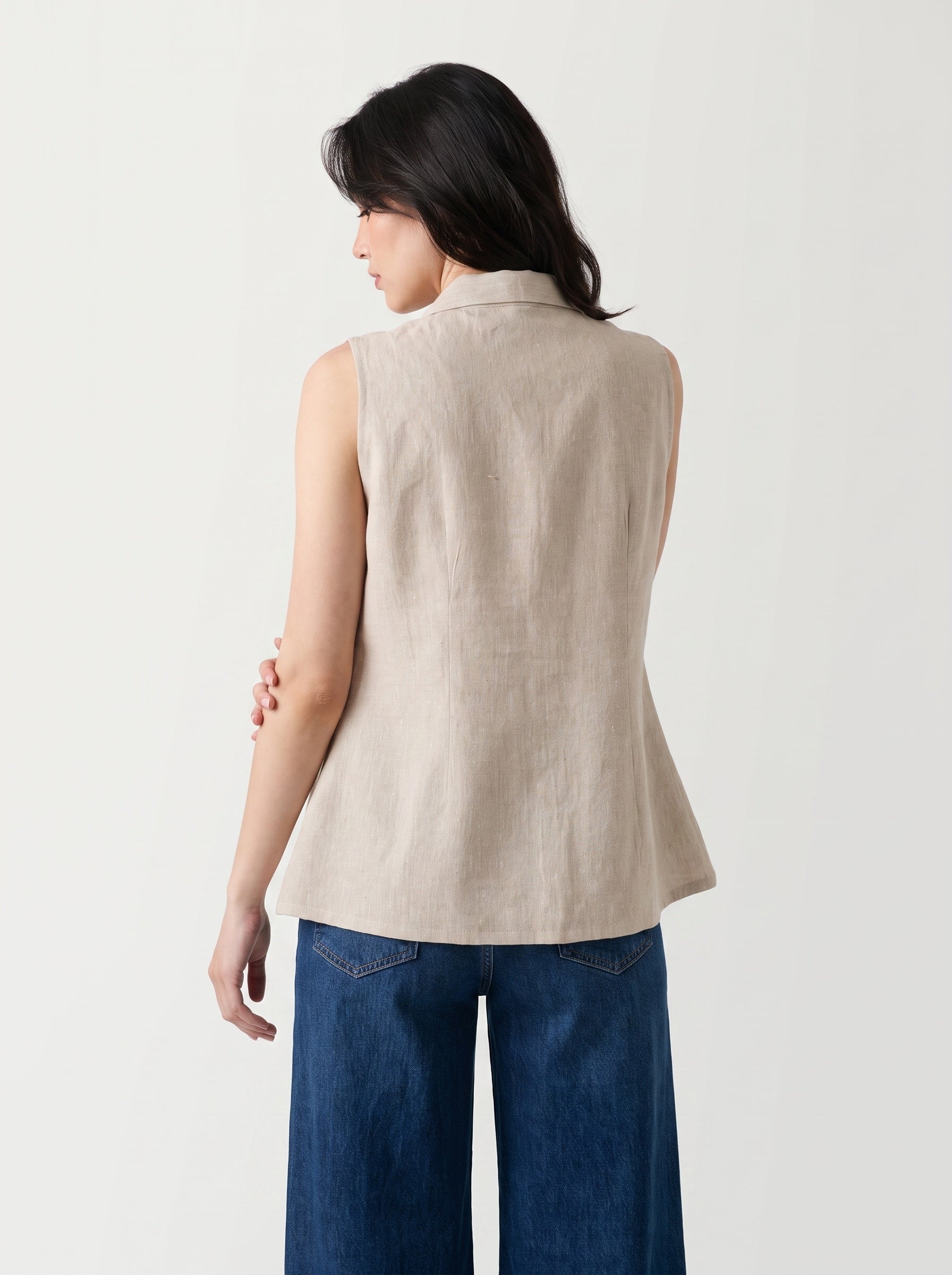 A woman shown from the back wearing a sleeveless beige linen vest and dark blue wide-leg jeans against a plain white studio background.