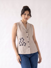 A woman modeling a sleeveless beige linen shirt with a scalloped collar, button front, and black floral embroidery on the front, paired with blue jeans against a plain white background.