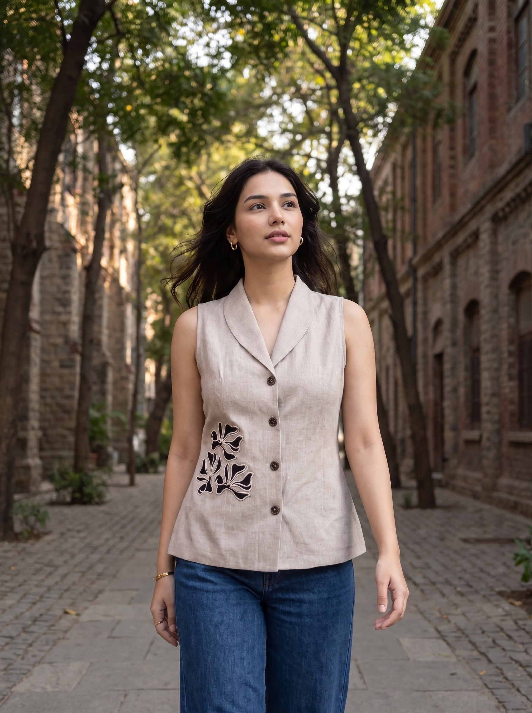 A woman stands on a cobblestone street lined with trees and brick buildings, wearing a sleeveless light beige linen vest with dark floral embroidery and buttons, paired with blue jeans.