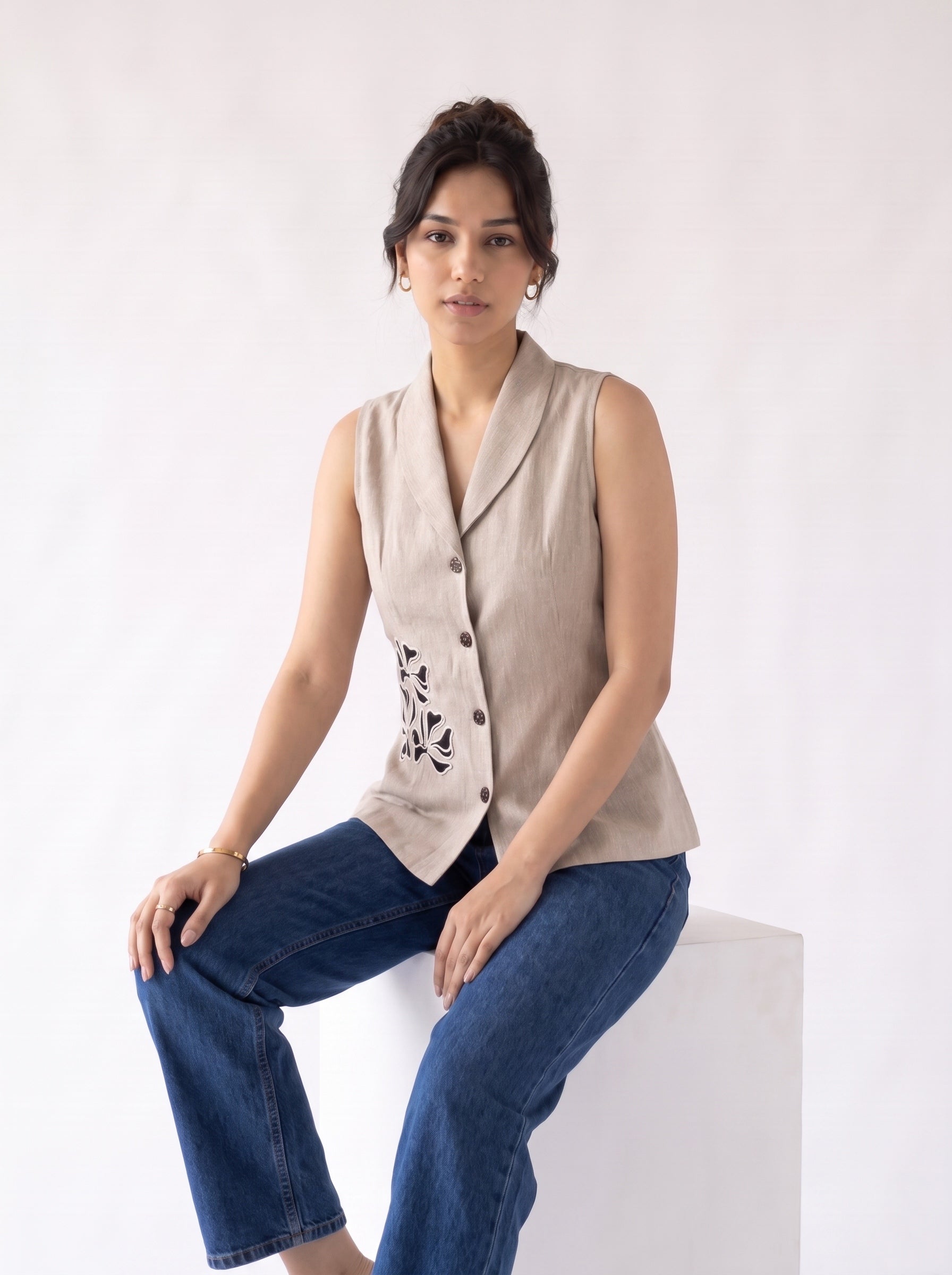 A woman sits on a white block against a plain white studio background, wearing a sleeveless beige linen vest with black floral embroidery and dark blue jeans.