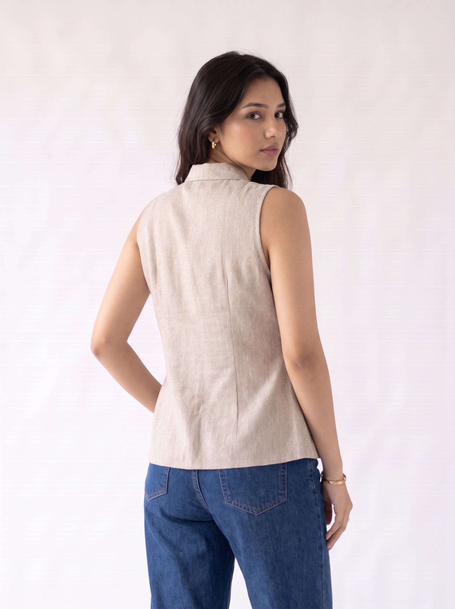 A woman wearing a sleeveless beige linen vest and blue jeans, photographed from the back against a plain white studio background.