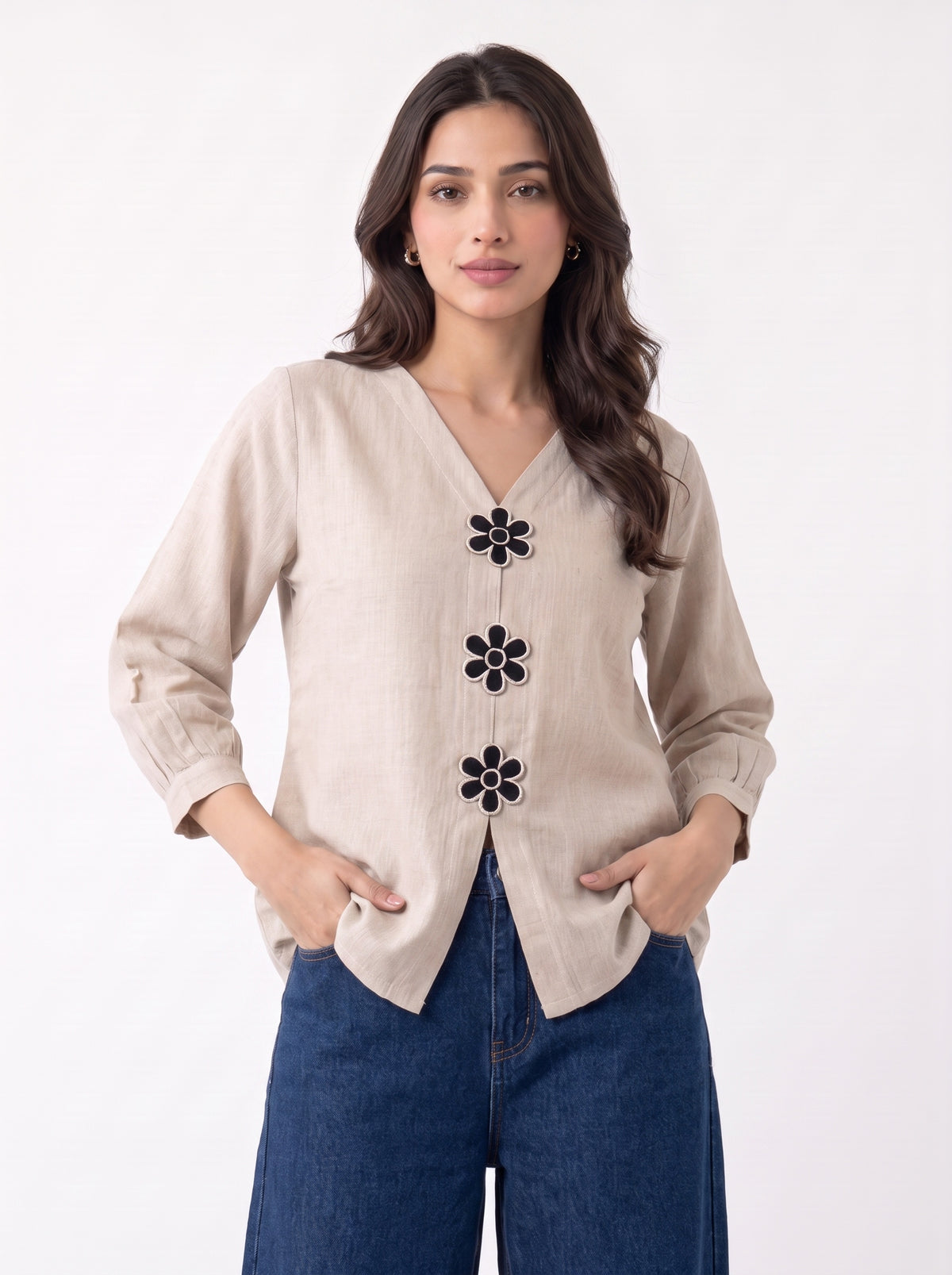 A woman modeling a beige linen-style cardigan with black embroidered flower details down the front, paired with blue jeans, against a plain white background.