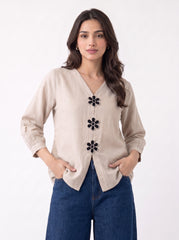 A woman modeling a beige linen-style cardigan with black embroidered flower details down the front, paired with blue jeans, against a plain white background.