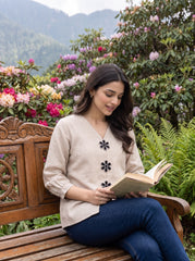 A woman sits on a wooden bench in a lush garden, reading an open book. She is wearing a light beige linen-blend top with black floral embroidery down the front and dark blue jeans.