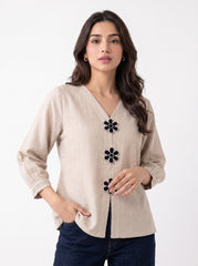A woman modeling a beige linen long-sleeve blouse with black and white flower-shaped buttons down the front, paired with dark blue jeans, against a plain white background.