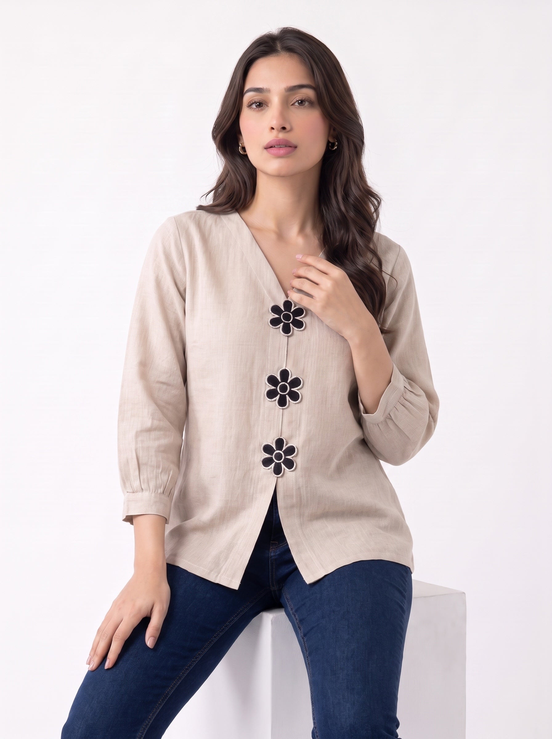 A woman wearing a beige linen-style cardigan with black flower-shaped buttons, paired with dark blue jeans, posing on a white block against a white studio background.