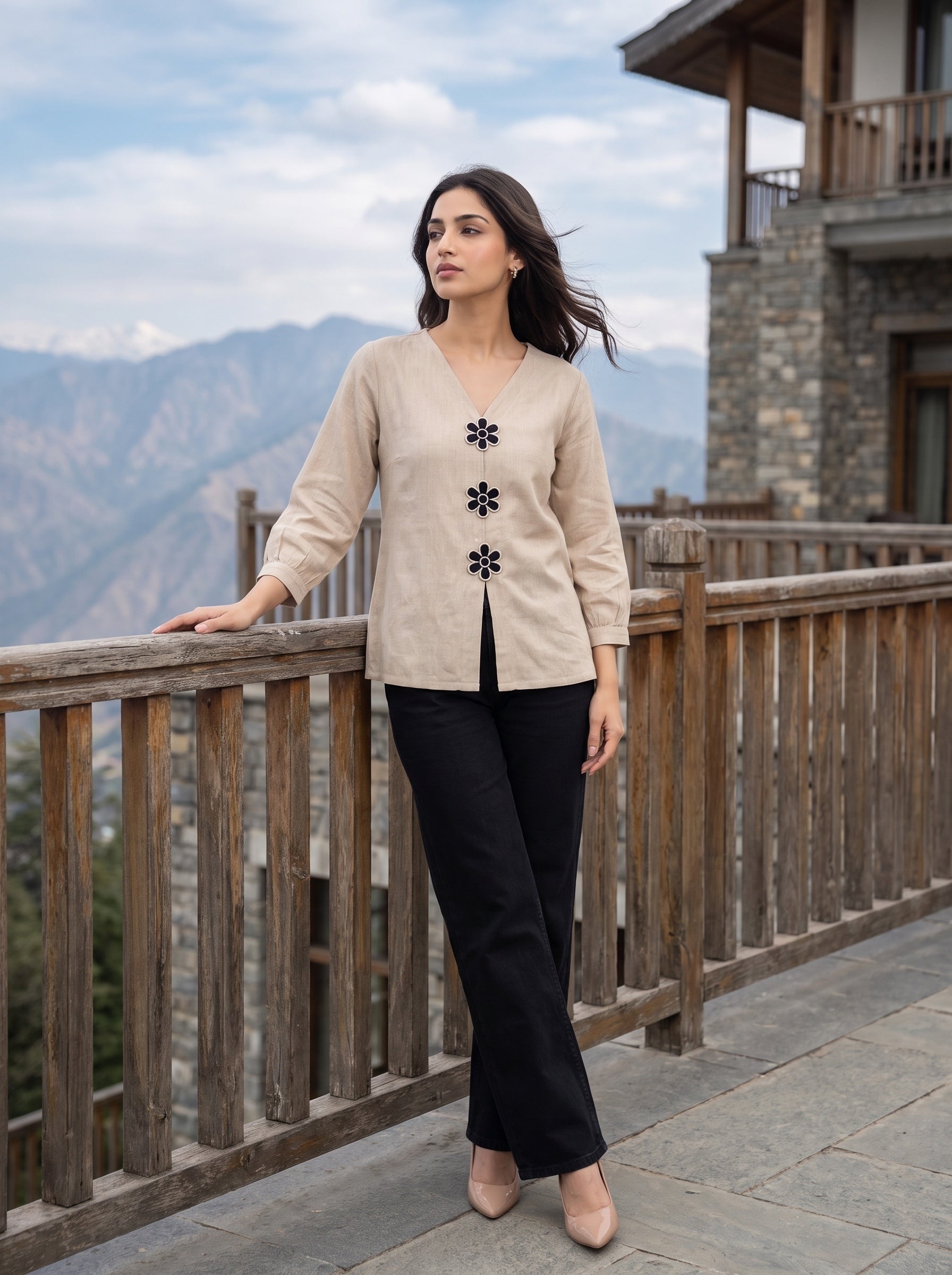 A woman stands on a stone walkway beside a wooden railing, wearing a beige long-sleeve cardigan with black decorative flower motifs down the front, paired with black pants and beige heels. Mountains and a stone building are visible in the background.