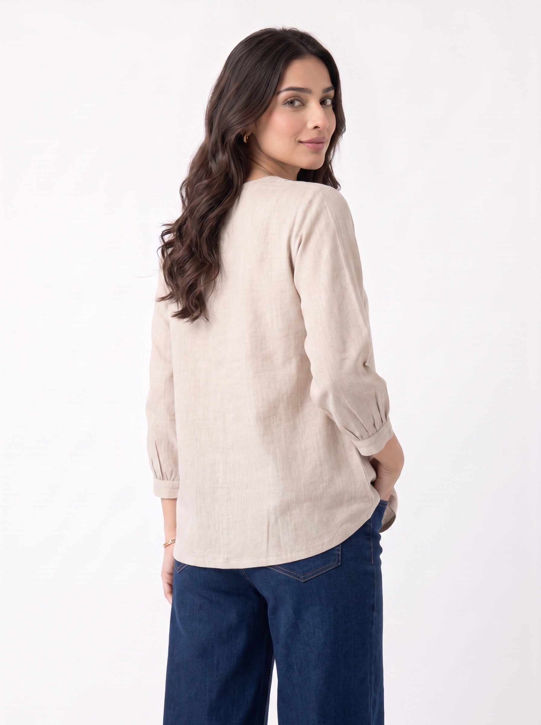 A woman with long dark wavy hair is shown from the back wearing a light beige linen long-sleeve top and dark blue jeans against a plain white background.