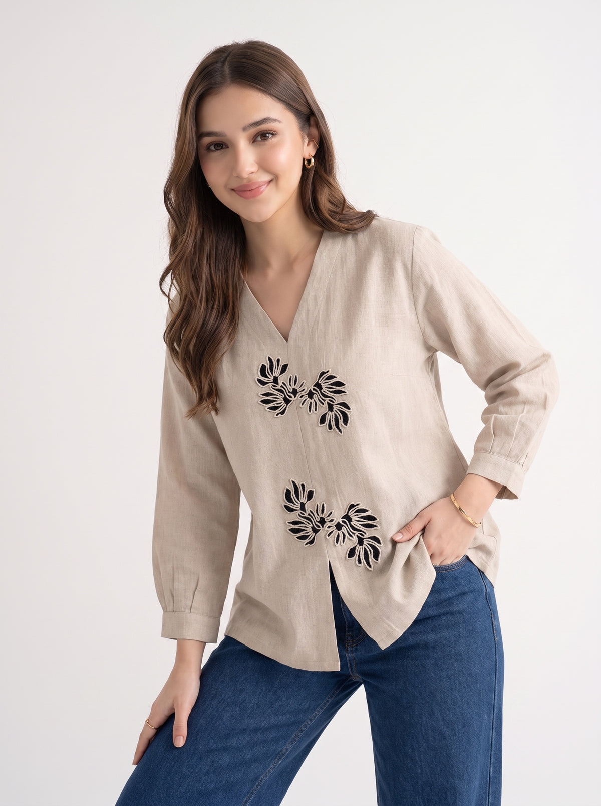A woman modeling a beige linen long-sleeve blouse with black floral embroidery on the front, paired with blue jeans, against a plain white studio background.