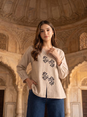 A woman wearing a beige long-sleeve linen top with black floral embroidery and blue jeans, standing in front of a warm, arched stone architectural background.