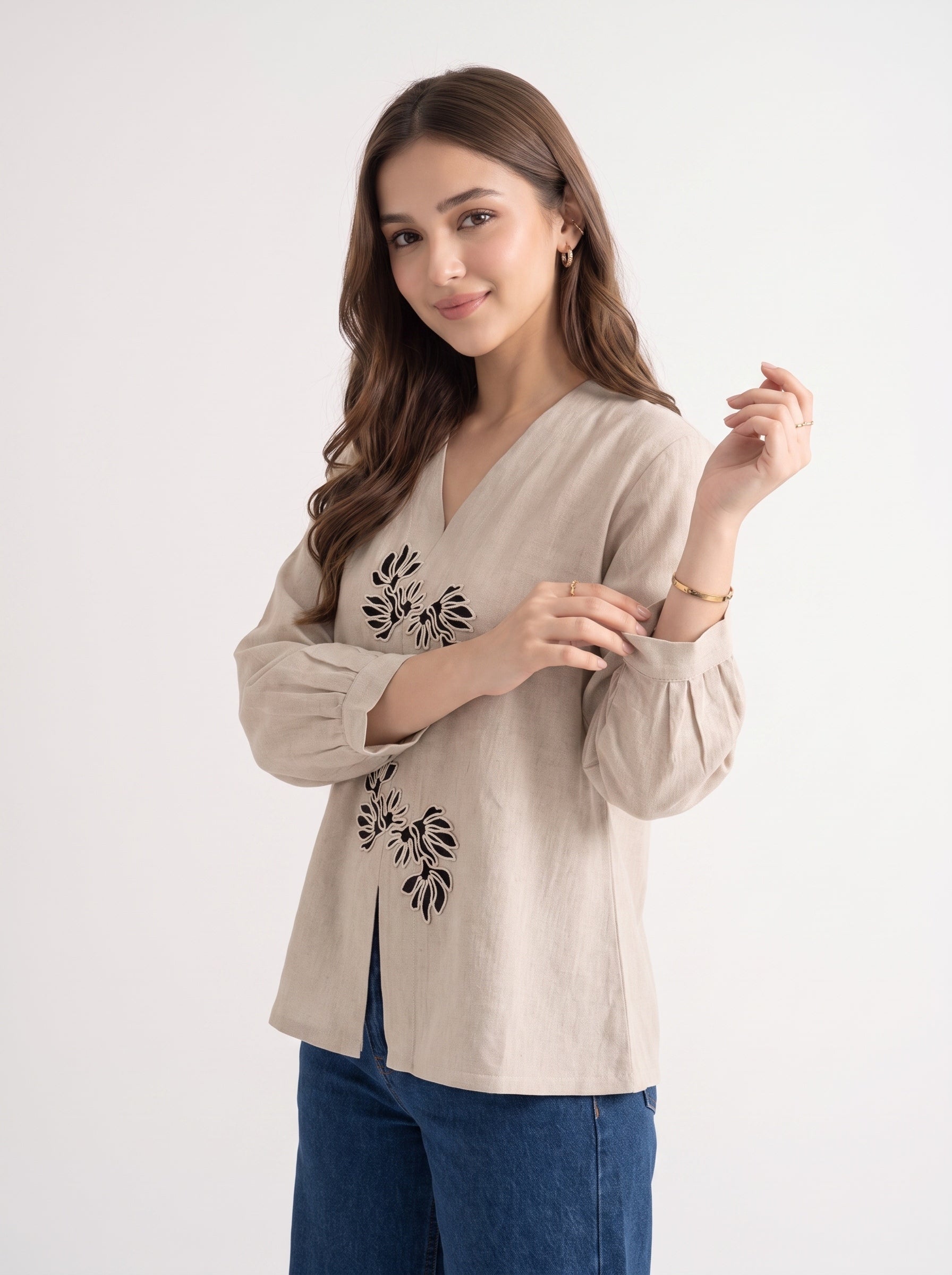 A woman modeling a beige linen-blend blouse with black floral embroidery on the front, paired with blue jeans, against a plain white studio background.