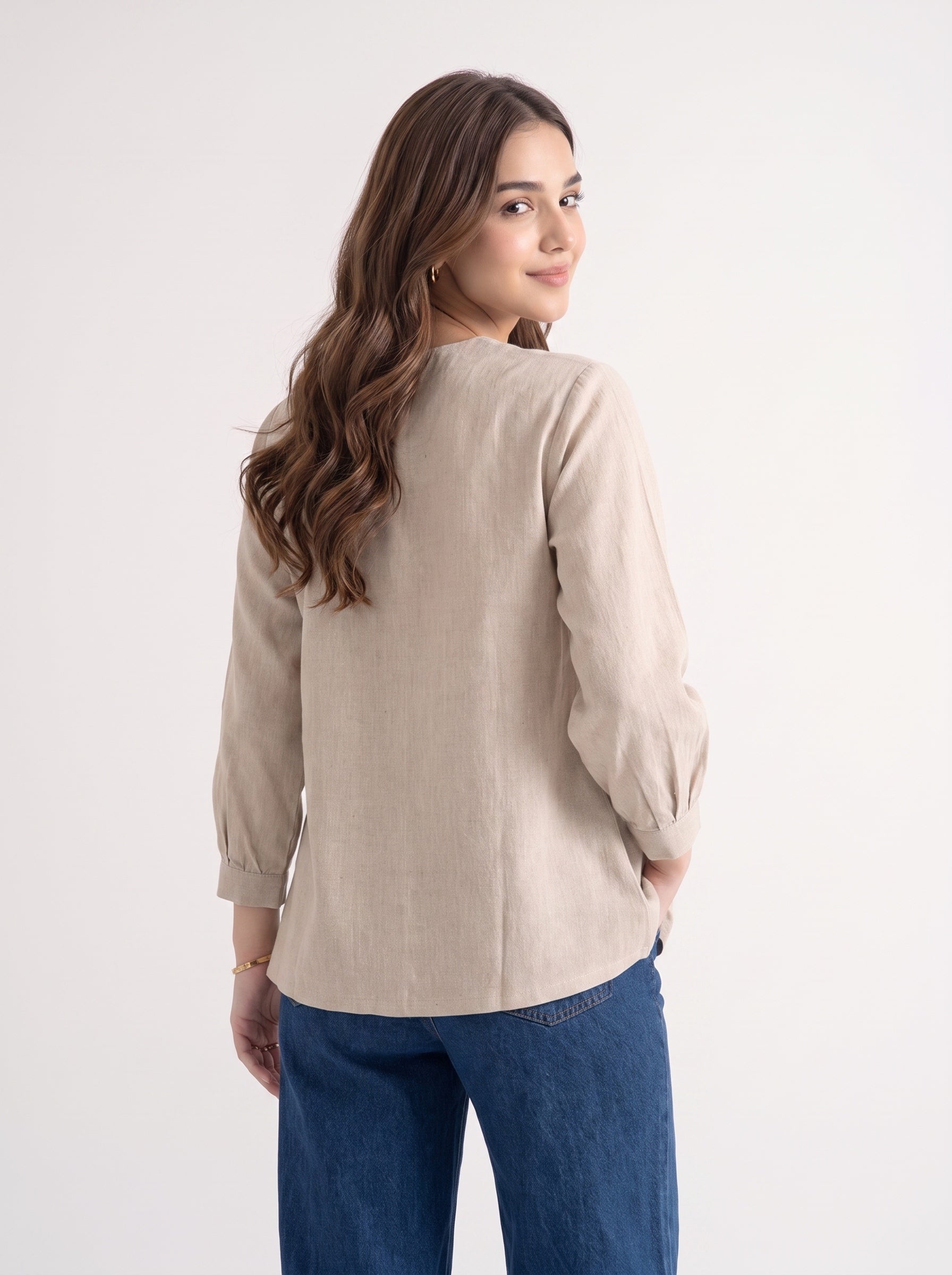 A woman with long brown hair is shown from the back wearing a light beige linen long-sleeve blouse with gathered cuffs and blue jeans, set against a plain white studio background.