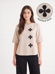 A woman modeling a beige short-sleeve top with three black floral appliques down the front, paired with black pants. A circular inset in the top right shows a close-up of the black-on-beige floral applique detail with the text 'Appliqué Work'.