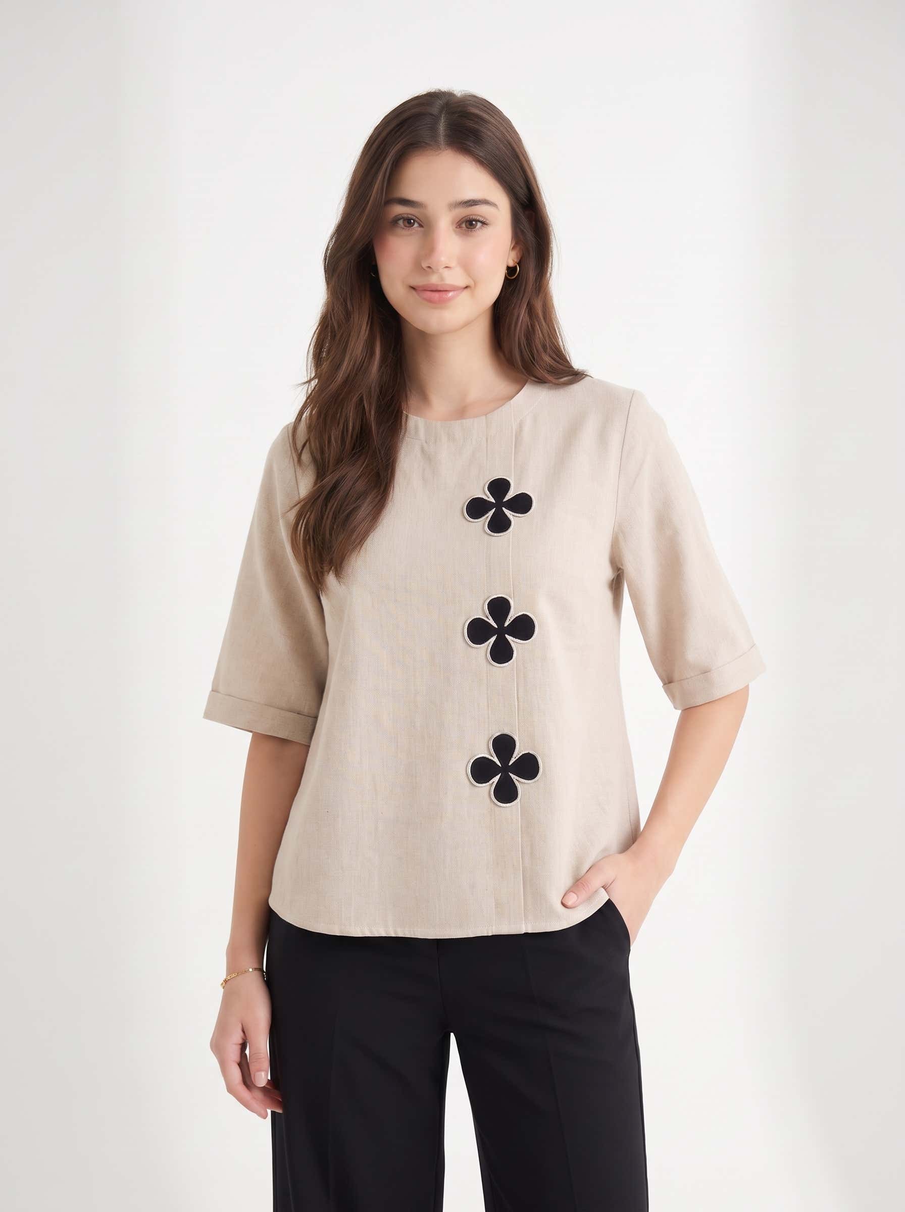 A woman modeling a beige short-sleeve top with three black embroidered flower motifs down the front, paired with black pants, against a plain light gray studio background.
