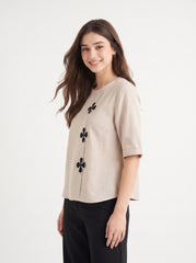 A woman modeling a light beige linen short-sleeve top with black embroidered clover motifs down the front, paired with black pants, against a plain white studio background.