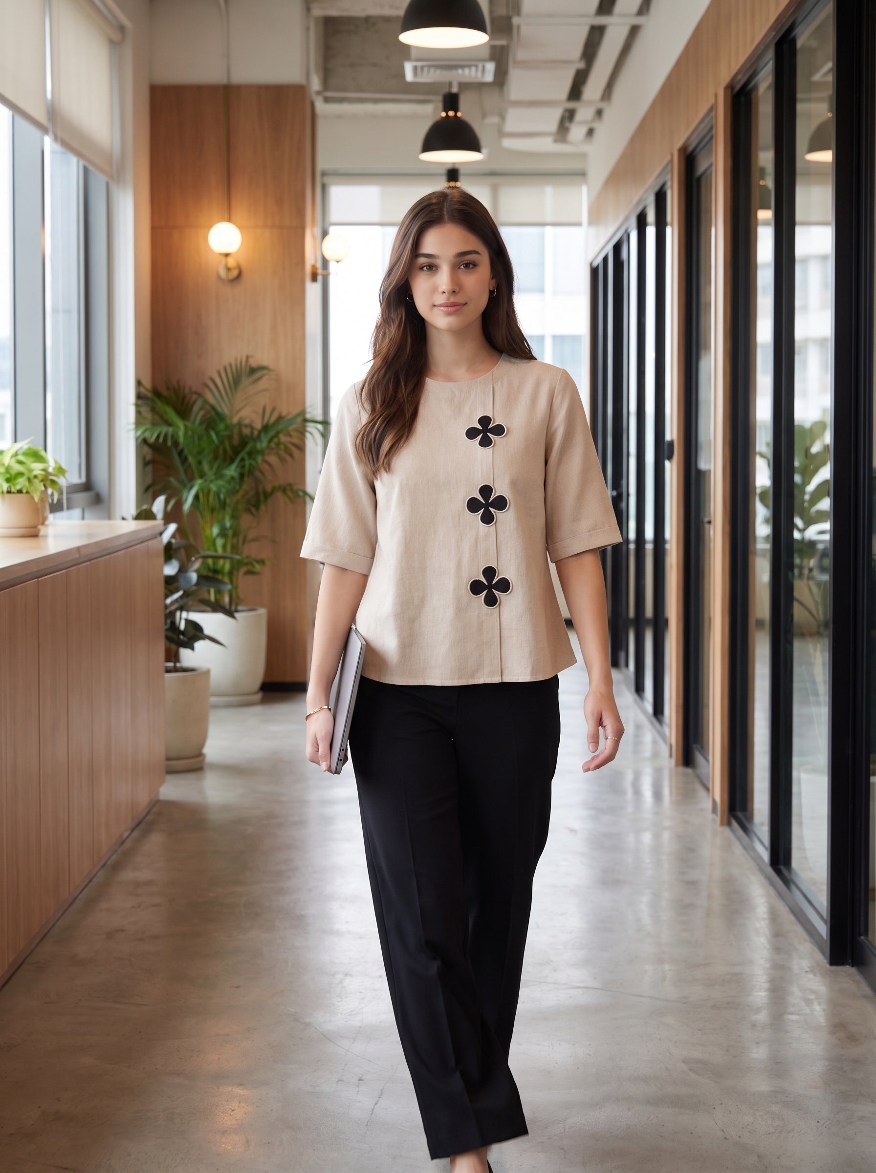 A woman walks down a modern office hallway wearing a beige short-sleeve blouse with black floral applique down the front, paired with black trousers.