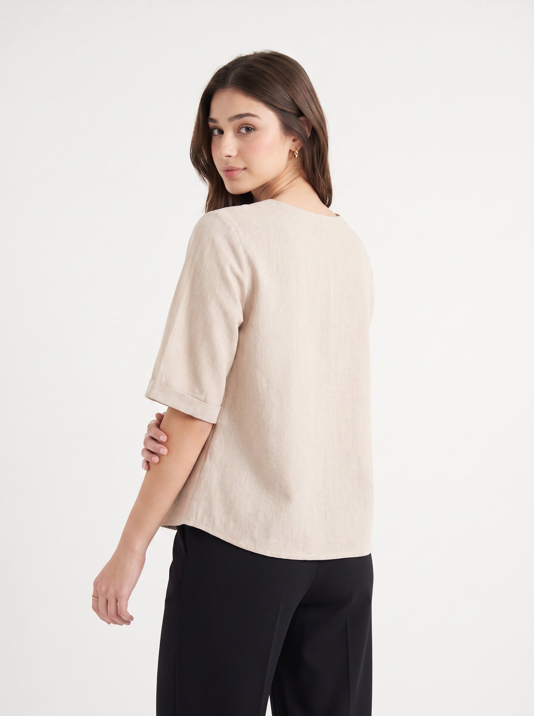 A woman wearing a light beige linen short-sleeve top with rolled cuffs and black trousers, photographed from the back against a clean white studio background.
