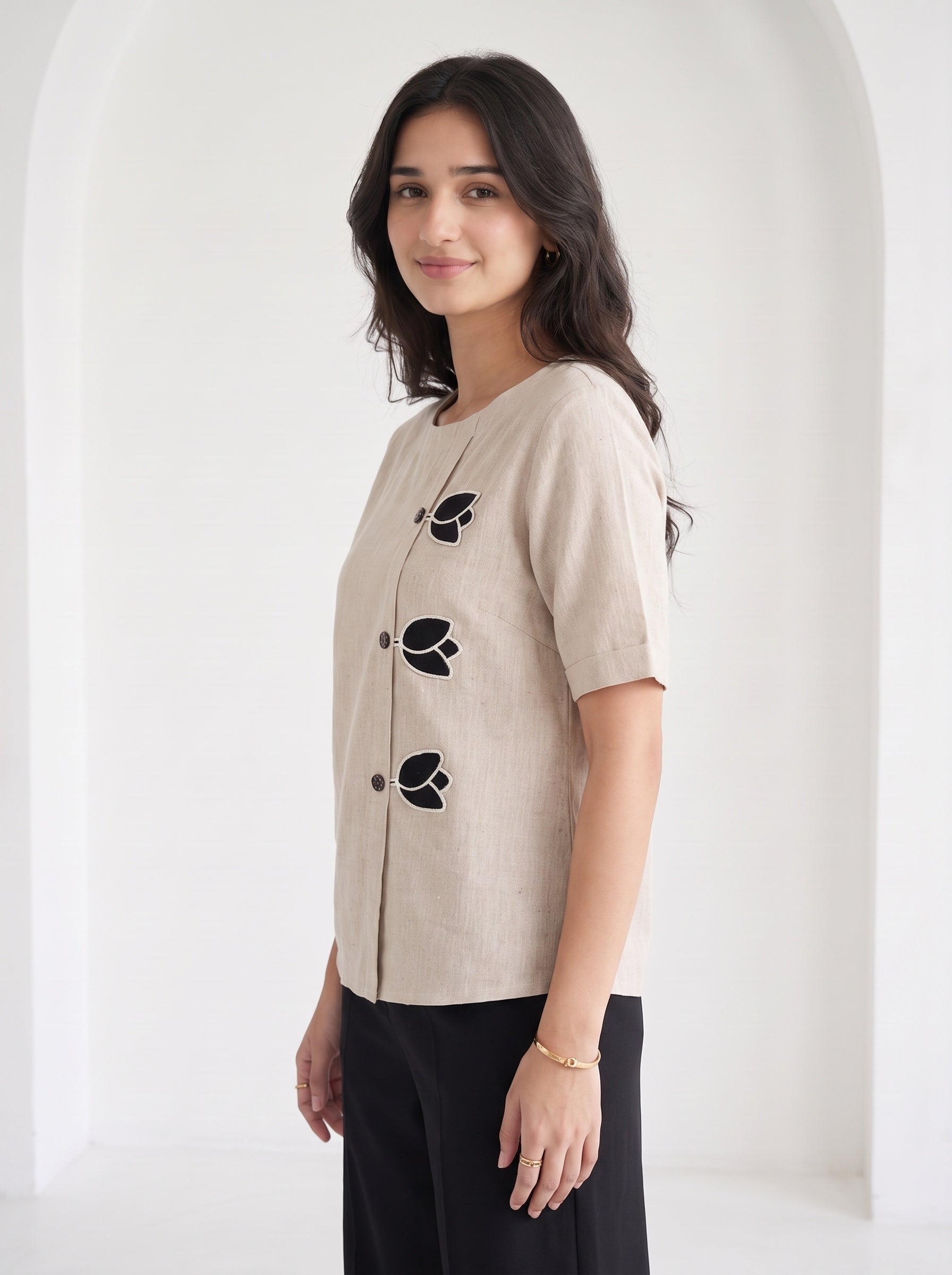 A woman wearing a beige linen short-sleeve top with black embroidered leaf details and black pants, standing in a bright white arched hallway.
