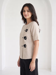 A woman wearing a beige linen short-sleeve top with black embroidered leaf details and black pants, standing in a bright white arched hallway.