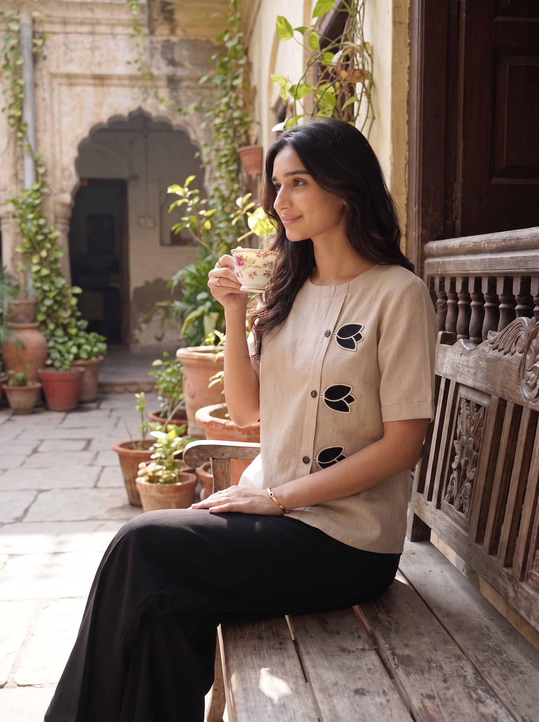 A woman sits on a wooden bench in a sunlit courtyard, holding a patterned teacup. She is wearing a beige short-sleeve top with black leaf embroidery down the front and black wide-leg trousers.
