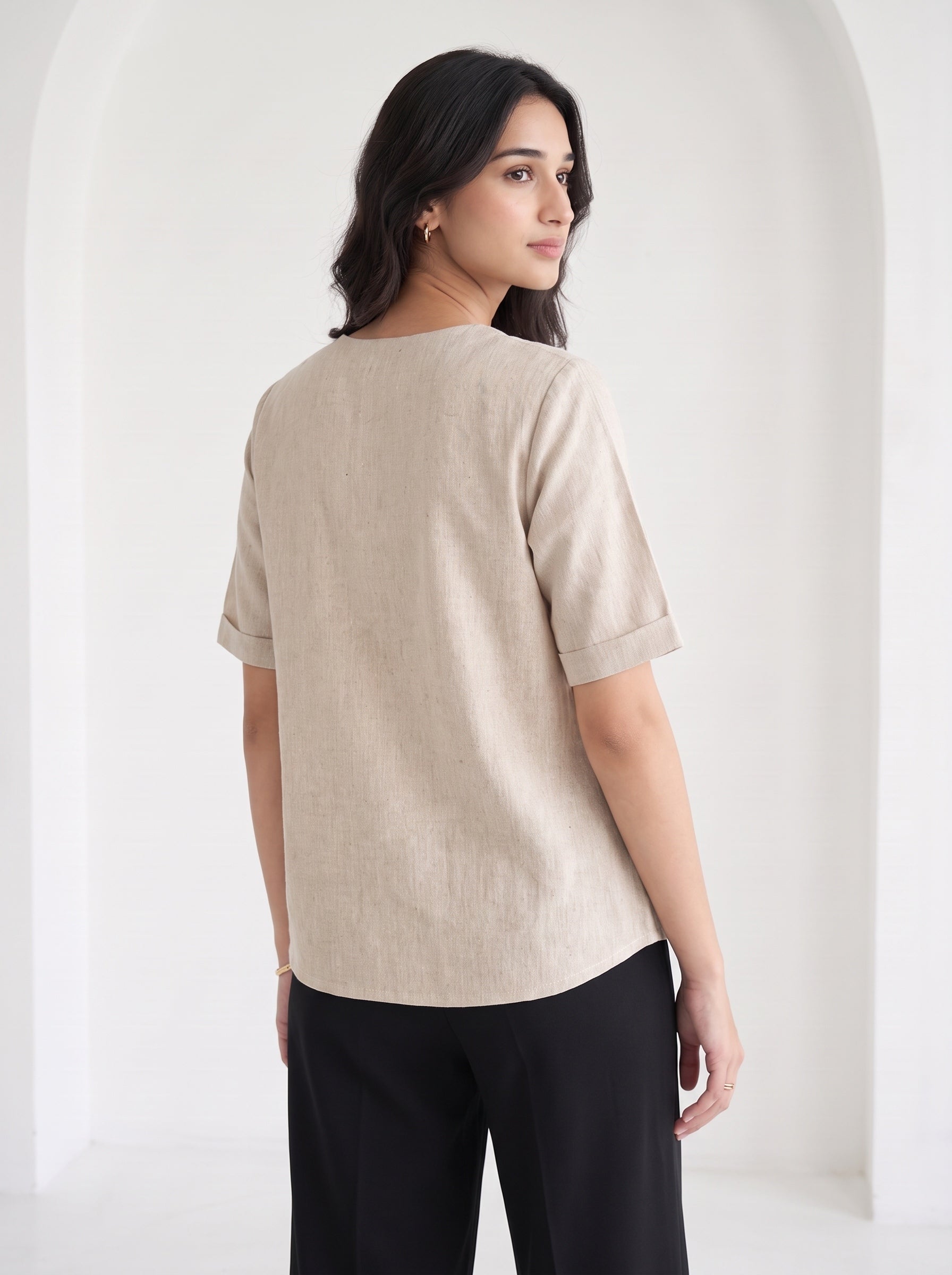 A woman shown from the back wearing a light beige linen short-sleeve top with rolled cuffs and black trousers, standing in a bright white arched interior.