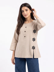 A woman modeling a beige linen tunic top with black embroidered flower details and a button placket, paired with blue jeans against a plain white background.