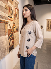 A woman wearing a beige linen three-quarter sleeve top with black embroidered flowers, paired with blue jeans, standing in a gallery next to a large woven wall hanging.